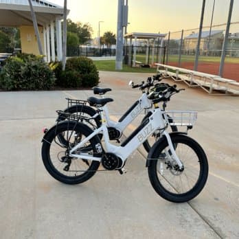 A white electric bike with a front basket and a rear rack is parked on a paved area near a tennis court, with greenery and a small building in the background.
